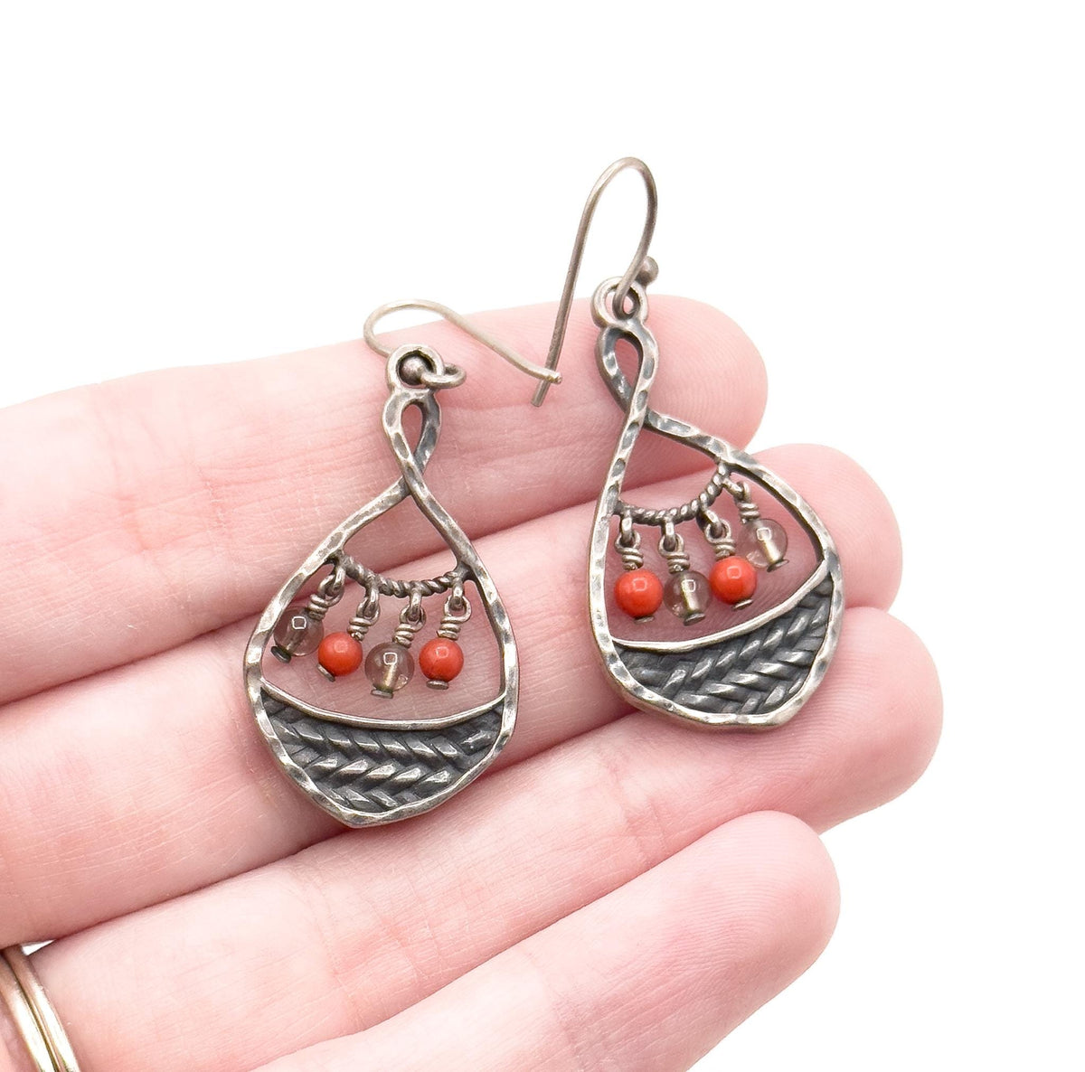 Both earrings resting across open fingers for scale, each hammered teardrop about as wide as a fingertip, ear wires angled outward above the dark braided panels and their small hanging beads.