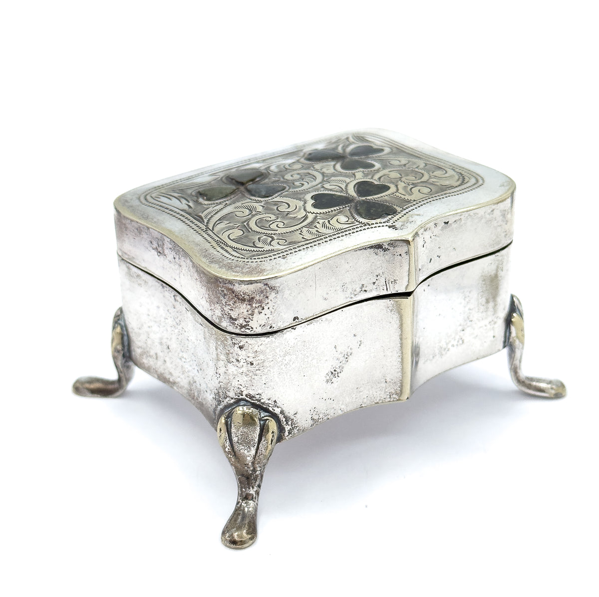 Low three-quarter view from a corner, the serpentine sides rising to the engraved lid with its clover insets while three teardrop feet splay out beneath the shaped base.