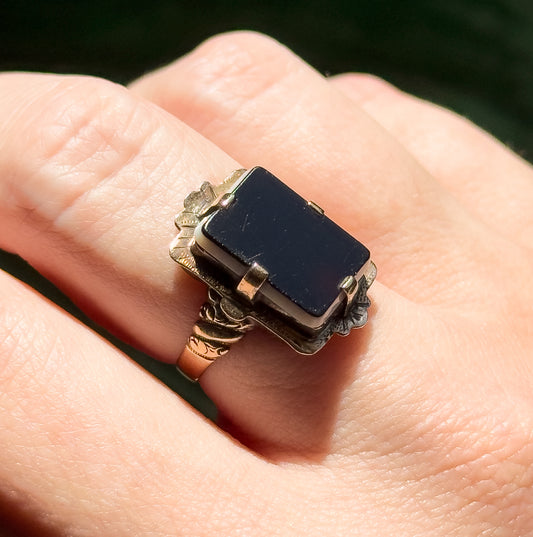 Shown on a finger in daylight, the rectangular sardonyx tablet and its prongs filling the finger's width, with the patterned shoulders and engraved shank visible alongside the skin for scale.