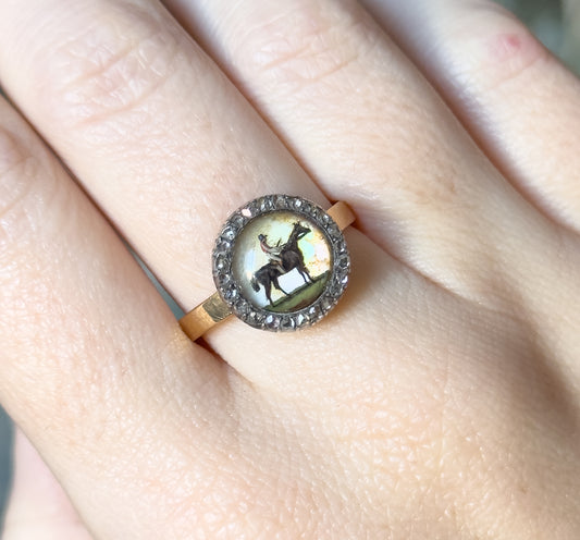Shown on a finger tilted toward the light, so the abalone backing flashes pale green and pink around the horse and rider while the flat gold shank crosses the finger below the bezel.