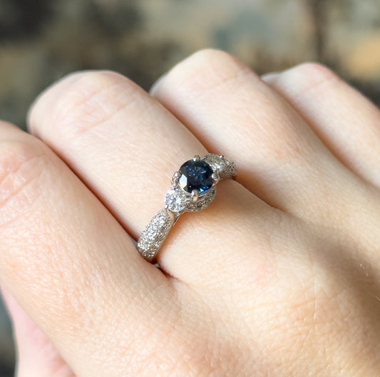 Shown on a finger at an angle, the platinum ring spanning about the width of the finger, with the blue sapphire basket, flanking diamond and pavé-set shoulders catching the light from above.