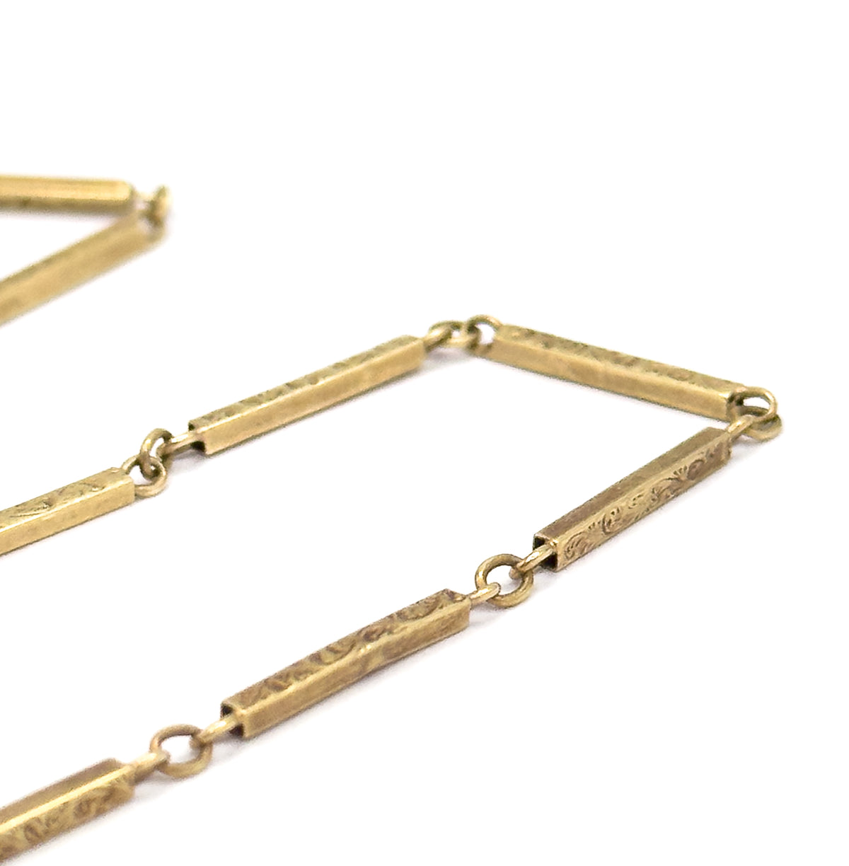 Angled close view of the chain zigzagging across white, the engraved scrollwork catching light along each bar face and the round jump rings looping through the squared link ends.
