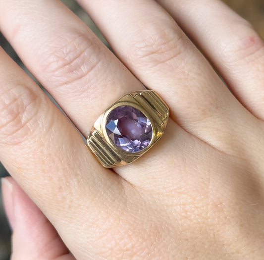 Closer view shown on the finger from above, the oval sapphire glinting pink-violet within its smooth bezel while the ridged shoulders step down on either side.