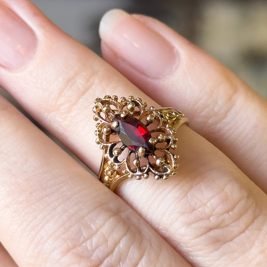 Shown on a finger at an angle, the openwork navette face fills the frame, its scrolled yellow gold loops and bead clusters framing the deep red marquise stone above the narrow shank.