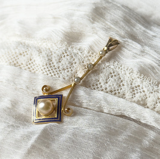 Art Deco pearl and seed pearl enamel lavalier pendant in 10K yellow gold, lying diagonally on white lace and gauzy fabric, its bail at top and cobalt blue enamel square at the lower end.