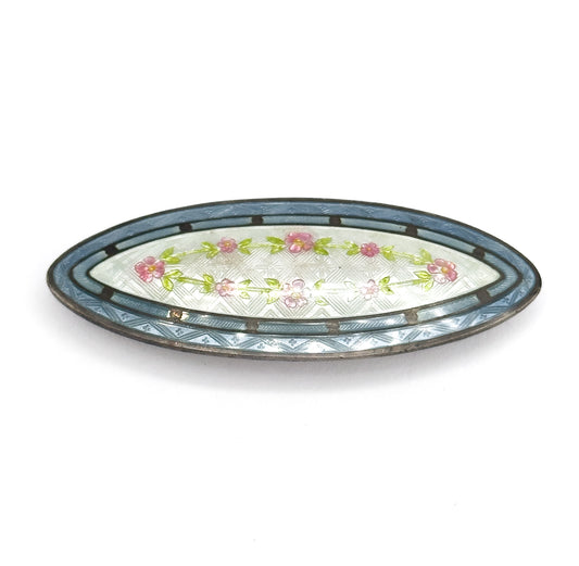 Low, tilted view of the same navette brooch on white, the frame's outer edge and the slight dome of the enamel visible, with the pink flower garland running the length of the cream centre.
