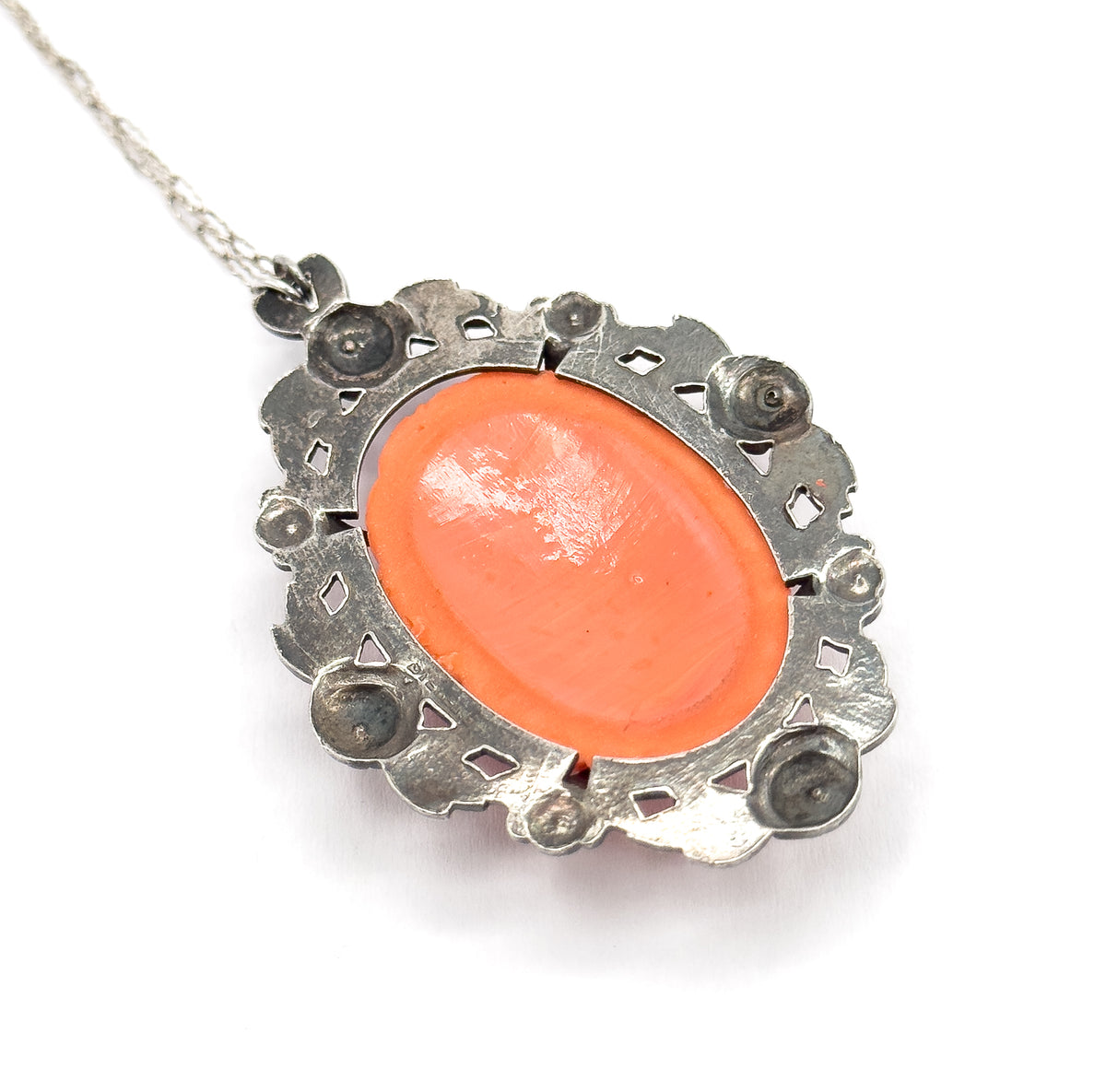 Reverse view showing the smooth back of the coral-coloured glass held in its bezel, the pierced silver frame behind it dotted with rounded settings, chain trailing off at upper left.