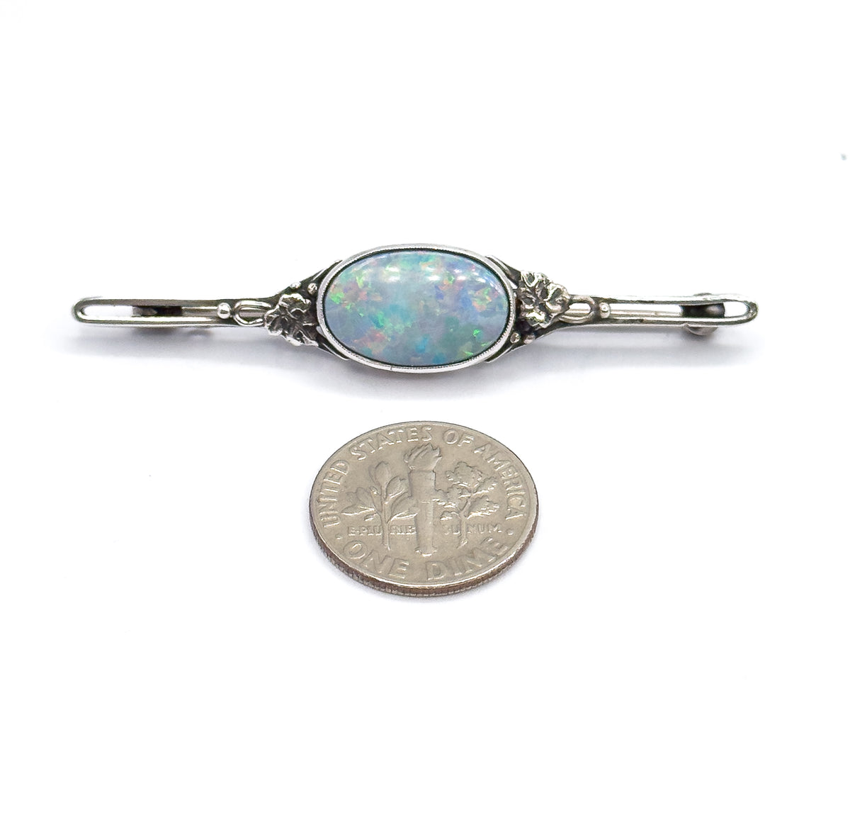 The brooch lies flat beside a dime for scale, its oval opal doublet in a bezel between floral silver mounts spanning a bar about three coin-widths long.