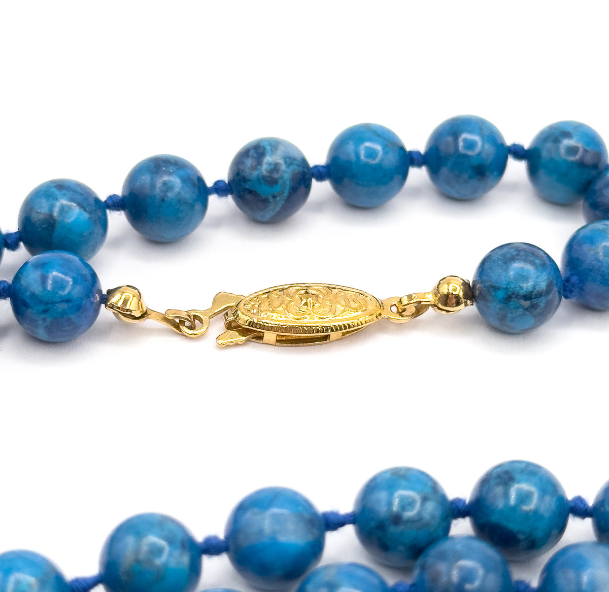 Close view of the oval yellow-toned clasp at the centre, its patterned face flanked by small round end caps and hooks that join the two rows of cobalt beads.