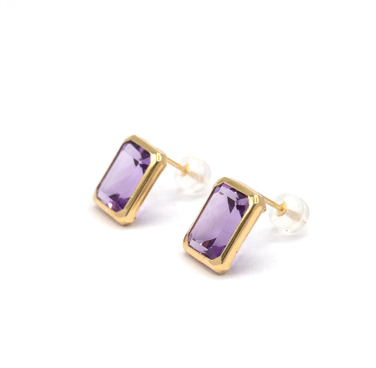 Both studs tipped slightly away so the yellow gold bezel rims read as a continuous polished frame around each soft purple emerald-cut amethyst, posts and clear backings trailing behind to the right.