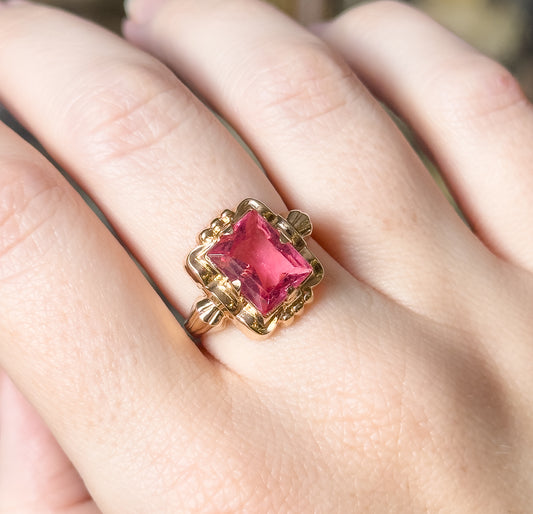 Shown on the hand at a closer angle, the pink glass stone catches light across its facets while the scalloped yellow gold flanks and shoulders sit low against the finger.