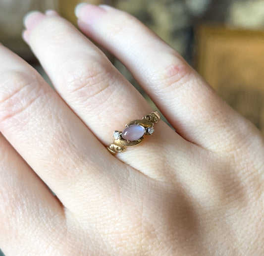 Shown on the hand from above, the ring sitting diagonally across the finger so its small scale is clear, with the pale blue-grey cabochon between the two diamonds in their prongs.
