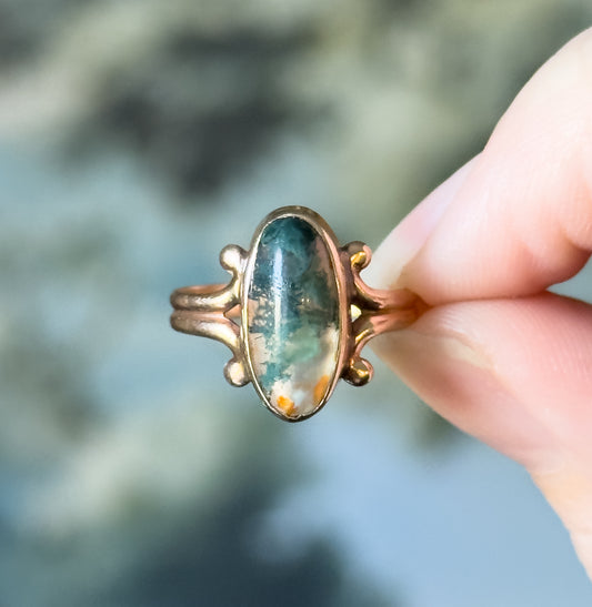 Edwardian moss agate cabochon ring in 10K yellow gold, held upright between two fingertips that show its small scale, the oval bezel-set stone flanked by scrolled shoulders.