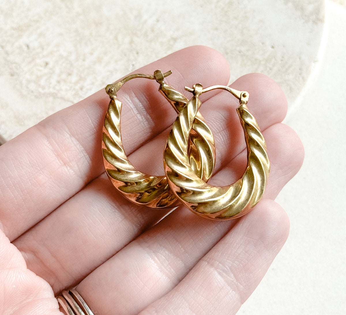 Both twisted gold hoops rested across a palm and fingers for scale, each about the length of a finger joint, with the hinged catch wires standing up at the top.