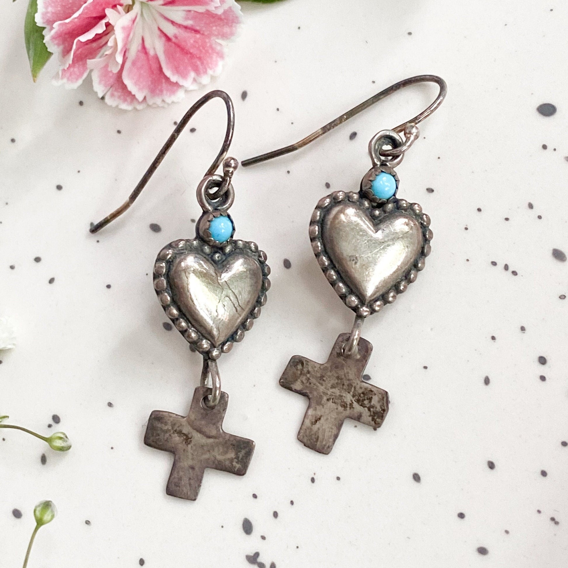 Heart-shaped earrings with cross details on a white background with pink flower and green leaves.