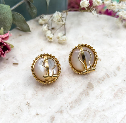 Straight-on rear view of both earrings, the hinged clip levers lying closed flat across the backs of the pearls inside their rope twist bezels, on the pale stone slab with dried flowers above.