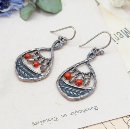 Pair lying at an angle on an old printed page with dried blue flowers above, showing the hook ear wires, hammered teardrop frames and braided silver band with red coral and clear glass beads.