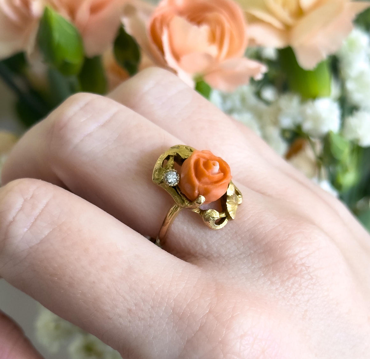 Shown on a finger against peach and white flowers, the ring face spanning about half the finger's width, with the coral rose sitting high beside the small bezel-set diamond and curling gold frame.