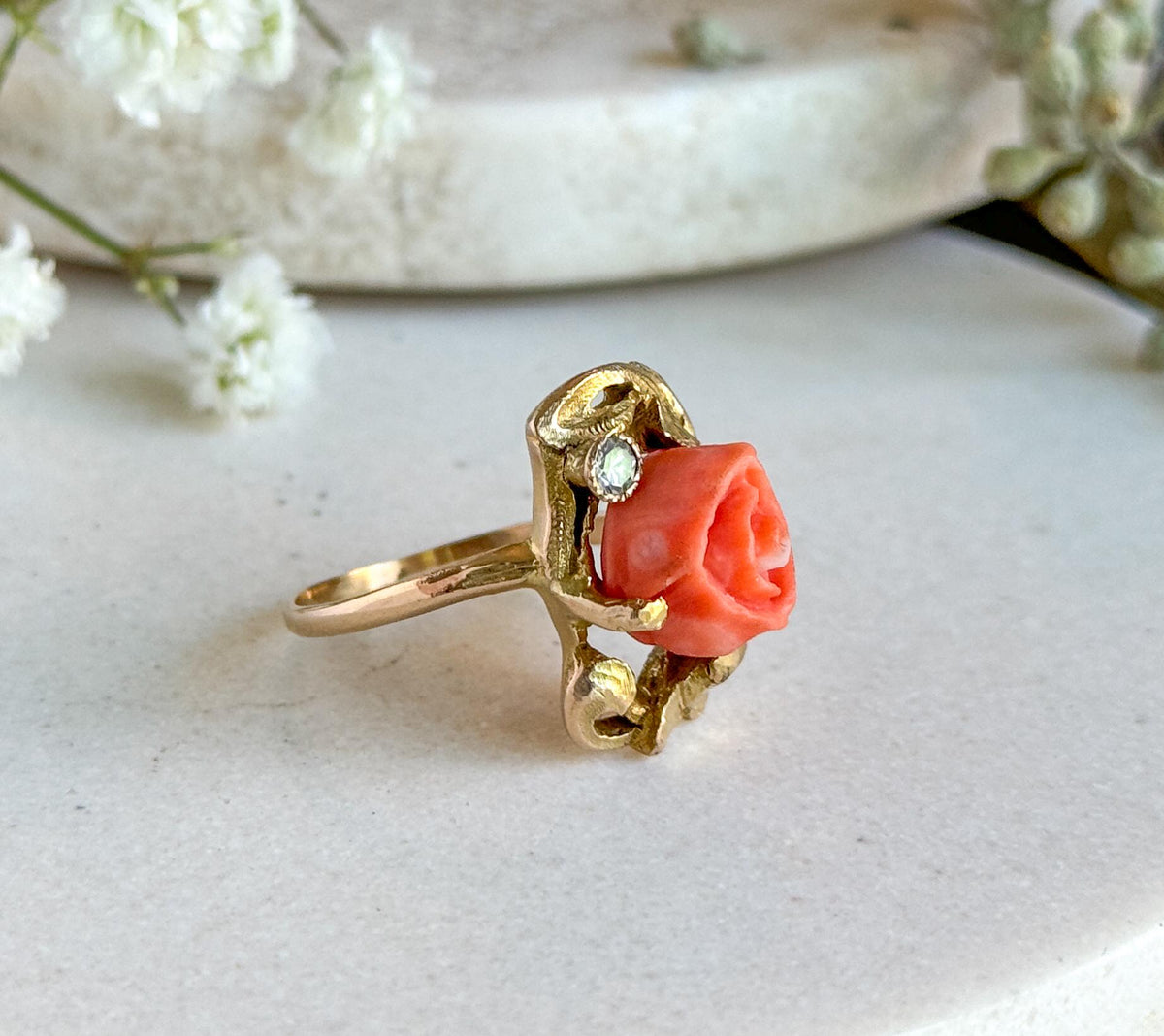 Side profile standing upright on a pale stone slab with sprigs of white blooms behind, showing the slim shank sweeping up to the raised coral rose and the small bezel-set diamond beside it.