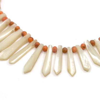A tight overhead crop of the centre curve, filling the frame with the longest mother of pearl pickets fanning downward and a single coral bead nestled between each flat drilled top.