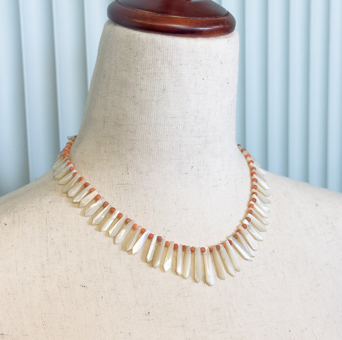 Edwardian mother of pearl and coral bead necklace shown at the neck on a linen dress form, its milky white drops fanning downward between small orange beads.