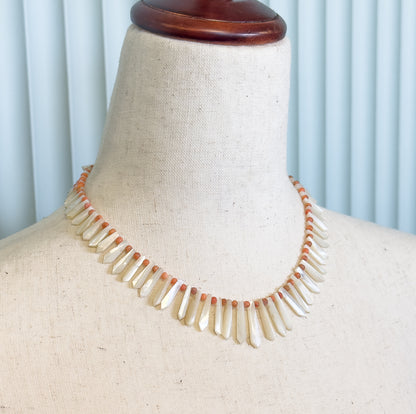 Edwardian mother of pearl and coral bead necklace shown at the neck on a linen dress form, its milky white drops fanning downward between small orange beads.