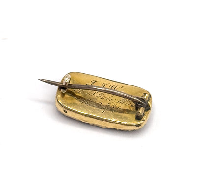 A tilted three-quarter view of the back, the engraved script running in lines across the panel while the pin stem crosses it diagonally between its coiled hinge and the hook of the catch.