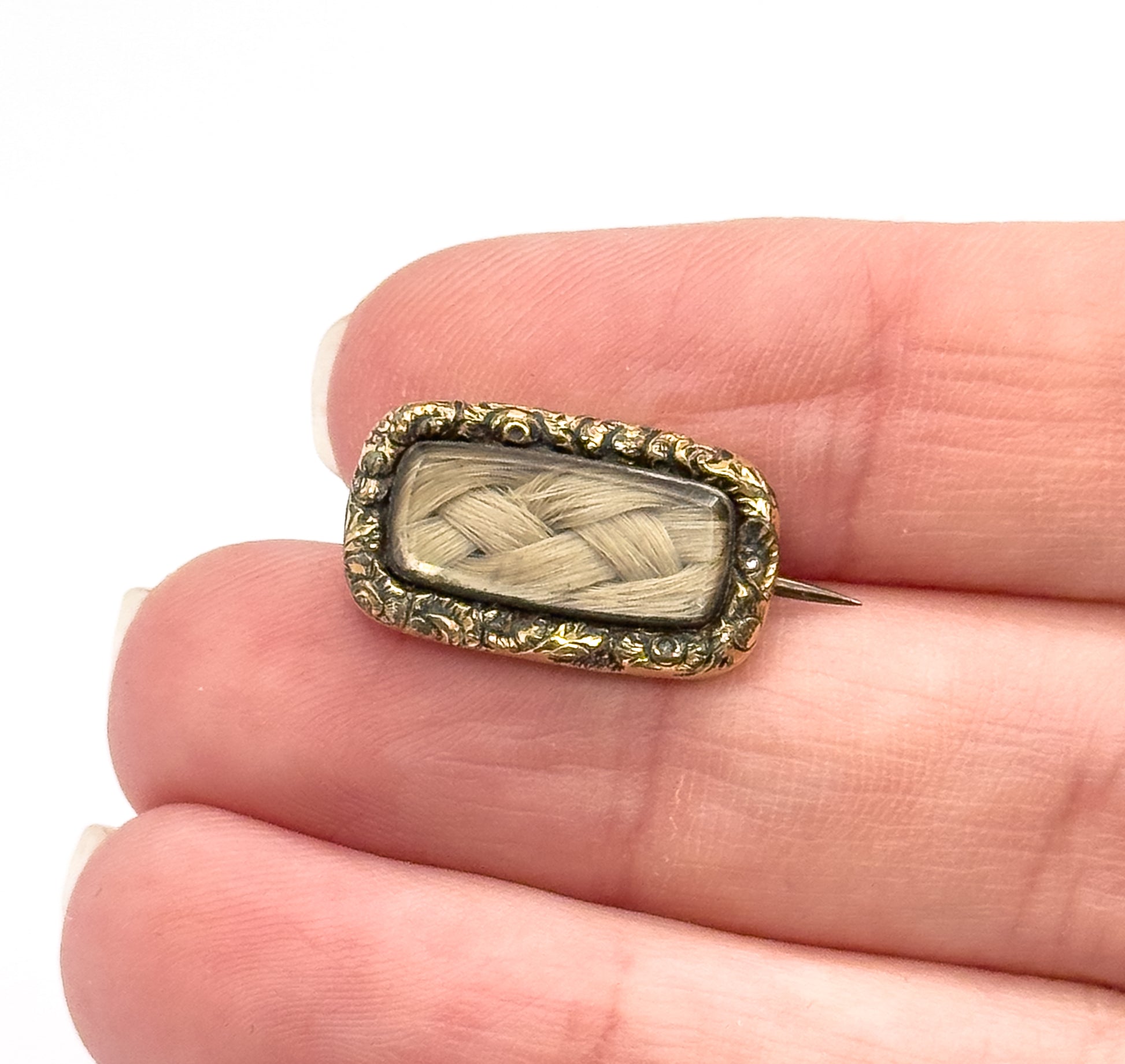 Resting on fingertips to show its small size, the front now faces up: woven blonde hair under glass inside a scrolling gold plated frame, with the pin stem just visible at one side.