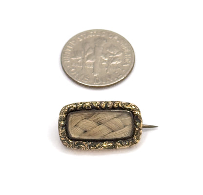 Beside a dime for scale, the brooch lies face up on white, the coin roughly matching the length of its woven blonde hair panel within the scrolling gold plated frame, pin stem angled out to one side.