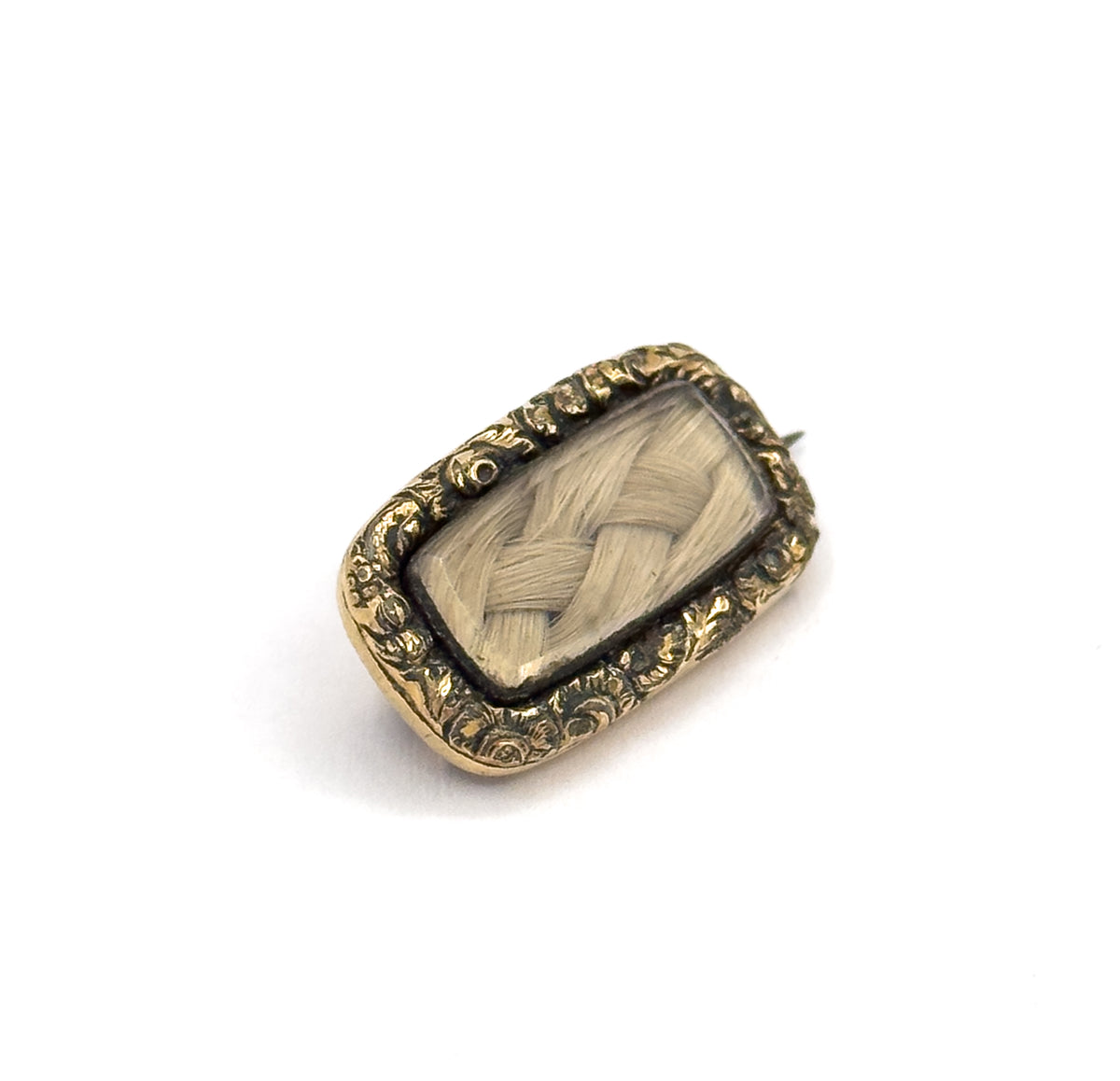 Tilted face-up close view on plain white, filling the frame so the criss-cross braid of blonde hair reads clearly under its glass, ringed by the scrolling floral gold plated frame, pin tip peeking out.