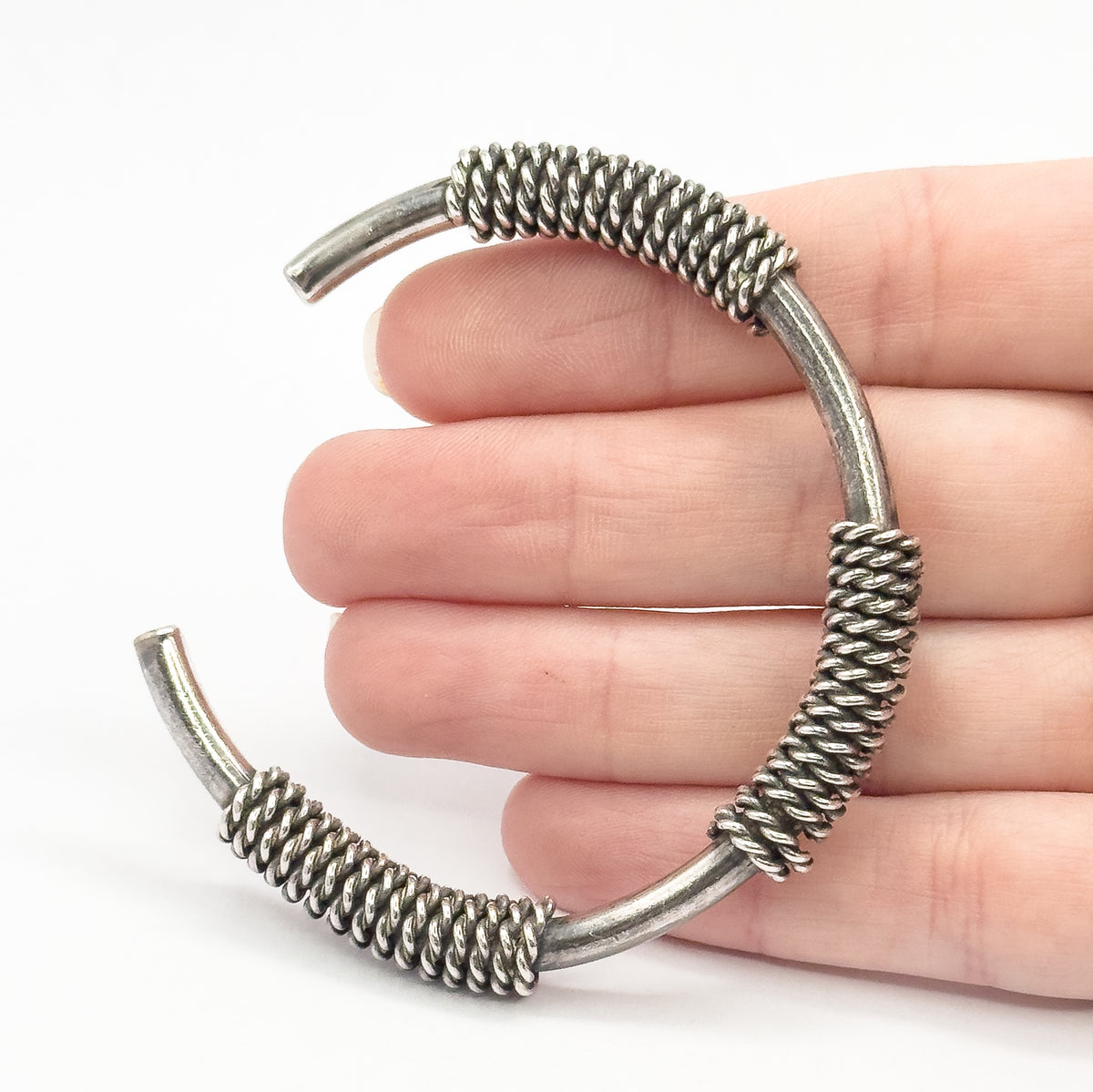 A sterling silver coiled cable cuff bracelet, its open ends curving apart, shown resting against the fingers of a hand for scale, with dark patina settling between the twisted coils.
