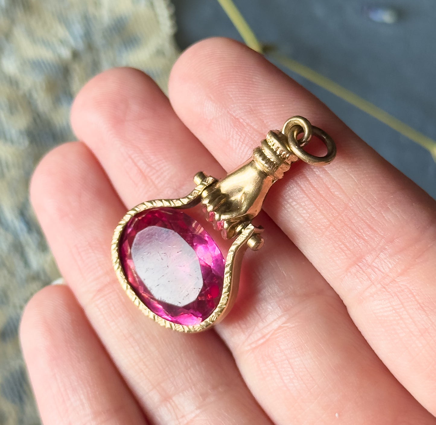 Resting flat across open fingers, which show its small scale, the hand fob lies with its round suspension ring at top and the oval pink glass gripped by the rosy metal fingers below.