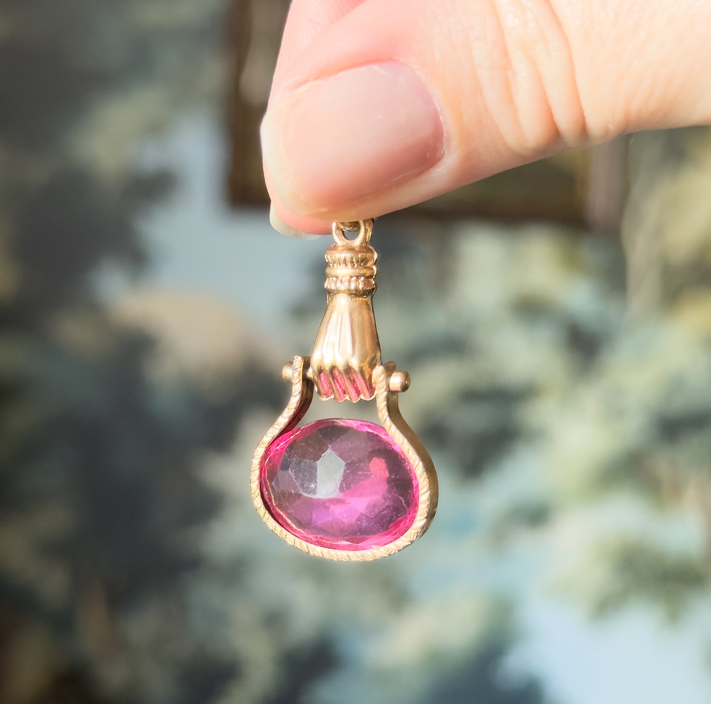 An Edwardian pink glass hand fob in base metal, held between fingertips against blurred greenery, its rosy metal hand gripping the oval faceted pink glass below a ribbed bail.