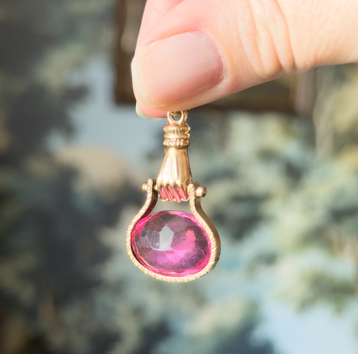 An Edwardian pink glass hand fob in base metal, held between fingertips against blurred greenery, its rosy metal hand gripping the oval faceted pink glass below a ribbed bail.