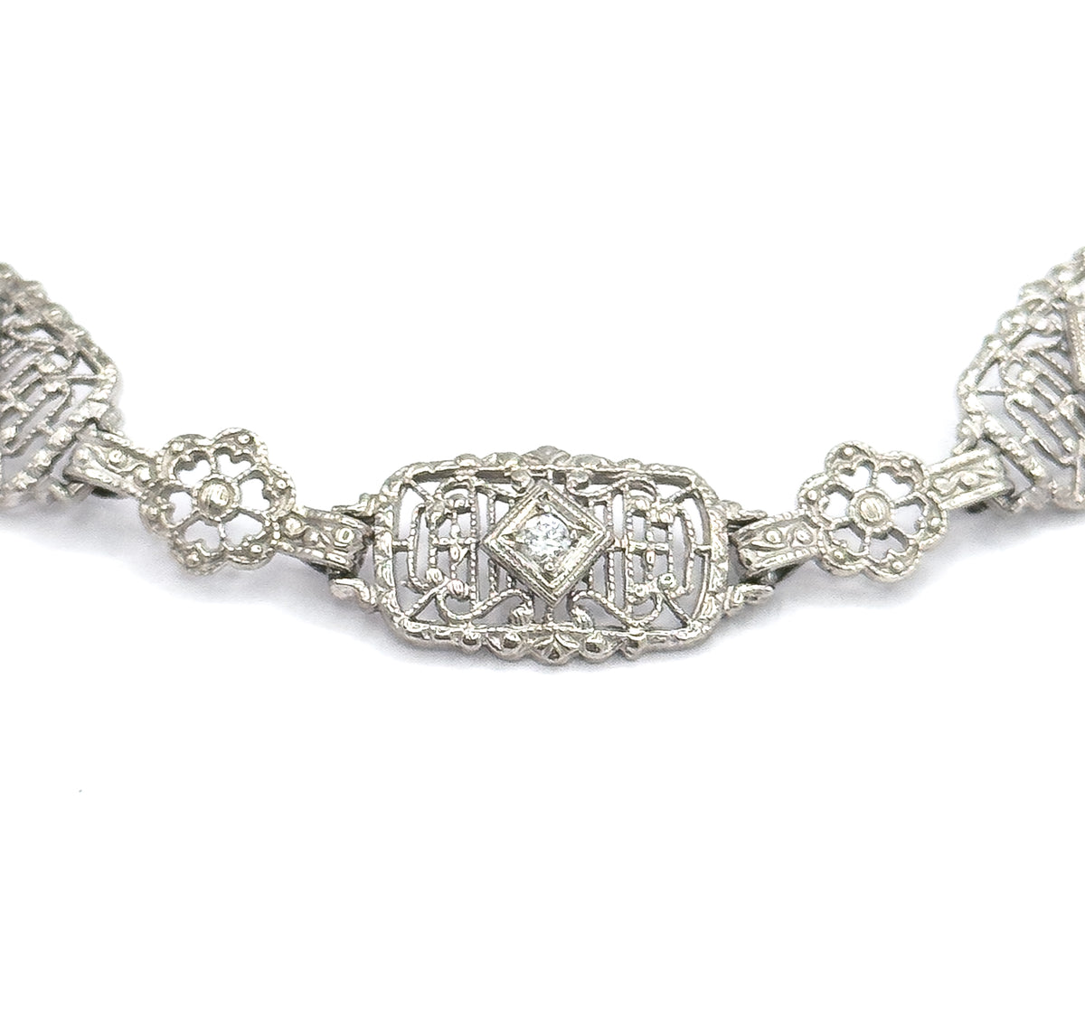 Straight-on macro of a single rectangular filigree link, its centre diamond-shaped setting holding one prong-set stone, with a four-petal floral link on each side and the next links cropped away.