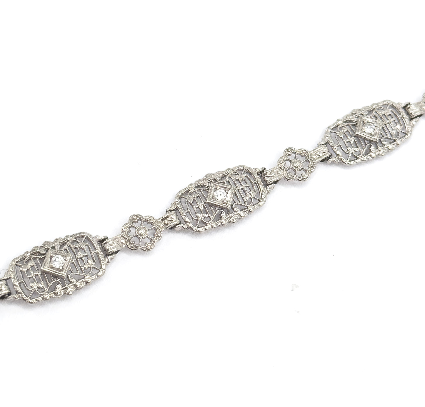 Three oval filigree links run diagonally across a plain white background, each centring a prong-set diamond in a diamond-shaped setting, with pierced floral links bridging between them.