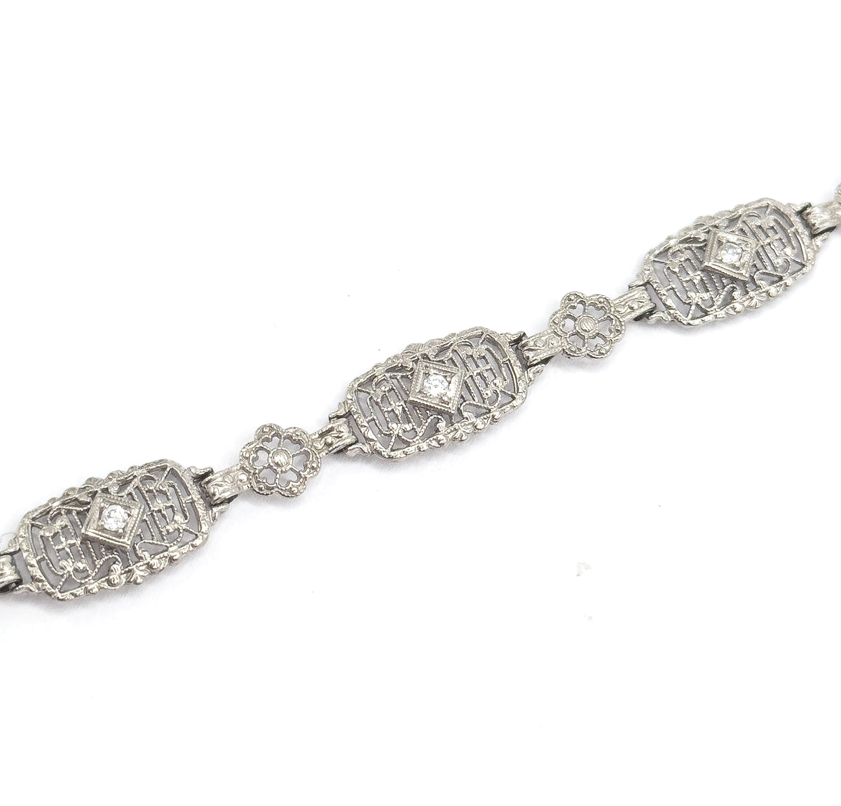 Three oval filigree links run diagonally across a plain white background, each centring a prong-set diamond in a diamond-shaped setting, with pierced floral links bridging between them.