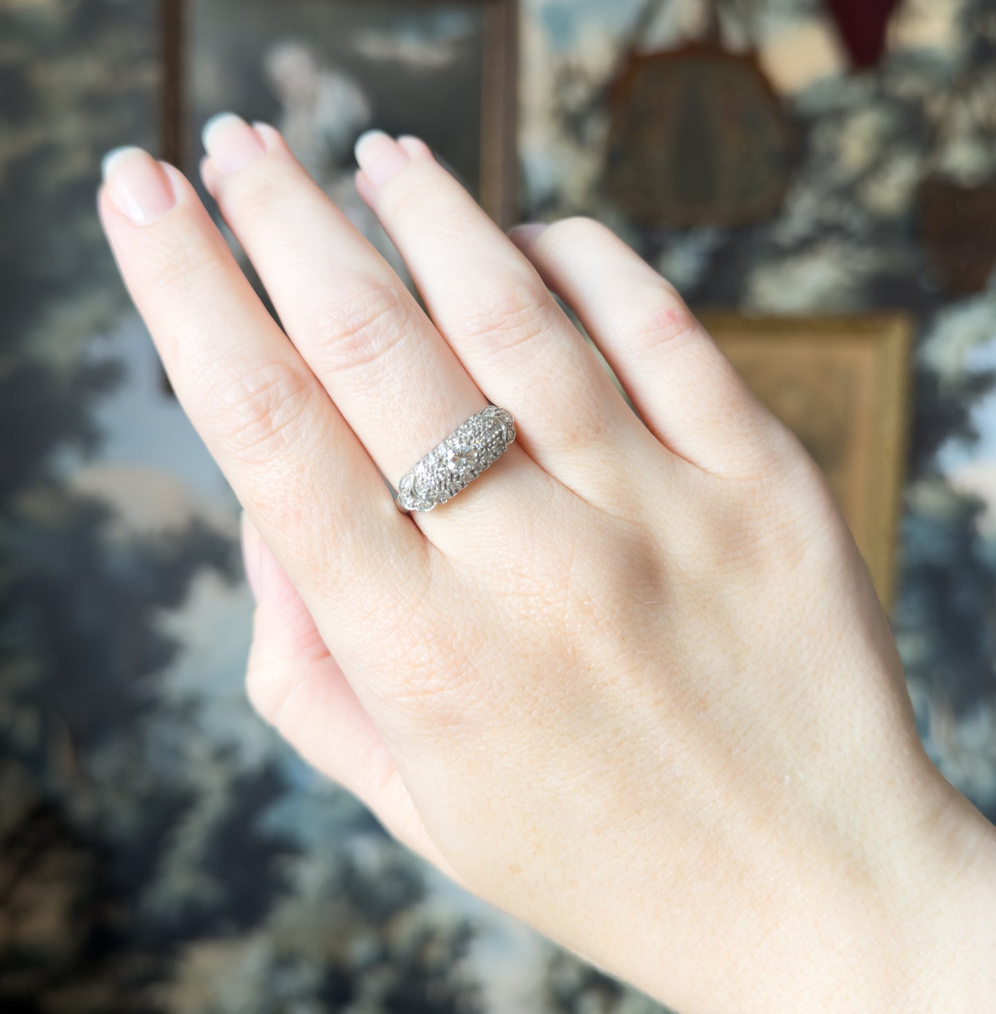The whole raised hand is in frame against a soft painted backdrop, the ring small on the ring finger, showing how the domed pavé face sits between the knuckles.