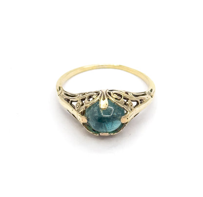 Tilted forward so the ring is seen from the top edge, the greenish blue cabochon sits low between prongs beneath the arching pierced fern gallery and slender split shank.