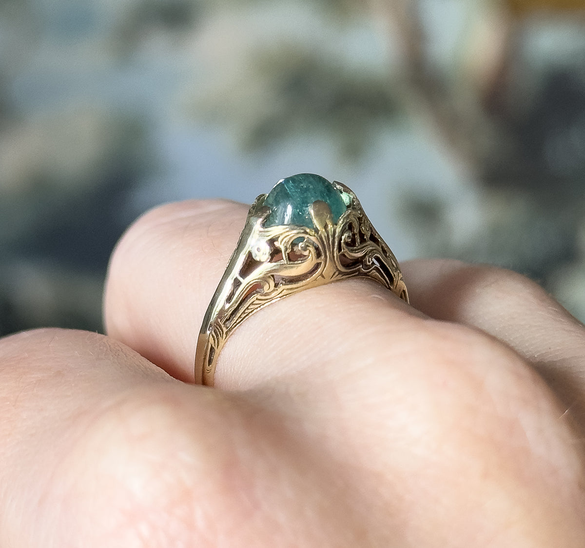 An Edwardian tourmaline cabochon ring in 14K yellow gold, shown on a finger, its pastel greenish blue oval stone prong-set above a pierced fern-like gallery and engraved shank.