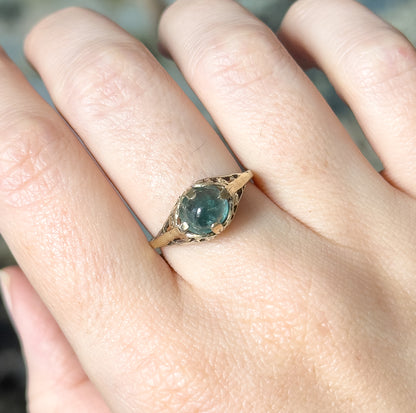 Close view from above of the ring shown on the finger, the greenish blue cabochon and its prongs catching light between the openwork shoulders that taper into the narrow shank.
