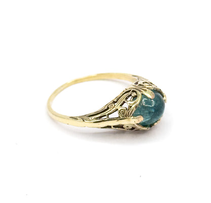 The ring off the hand against white, angled to show the pierced fern-like gallery from the side with the greenish blue cabochon held by prongs above the tapering engraved shank.