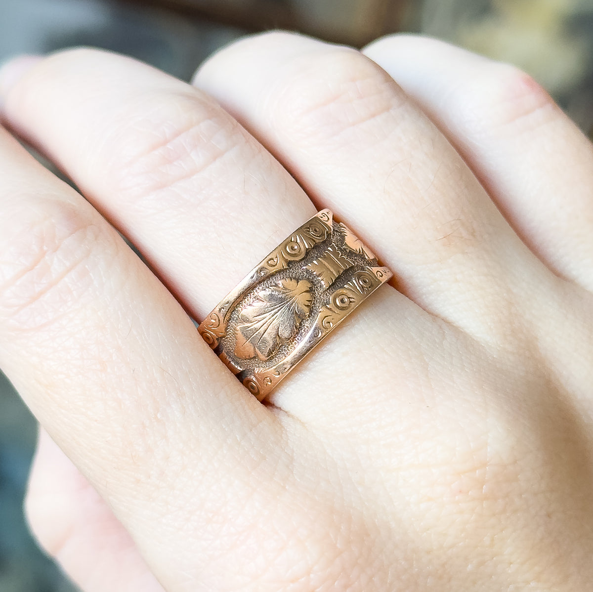 Close view with the hand tilted, the ring shown on a finger between the knuckles so the deeply cut leaf and scroll engraving fills the band's flat top face.