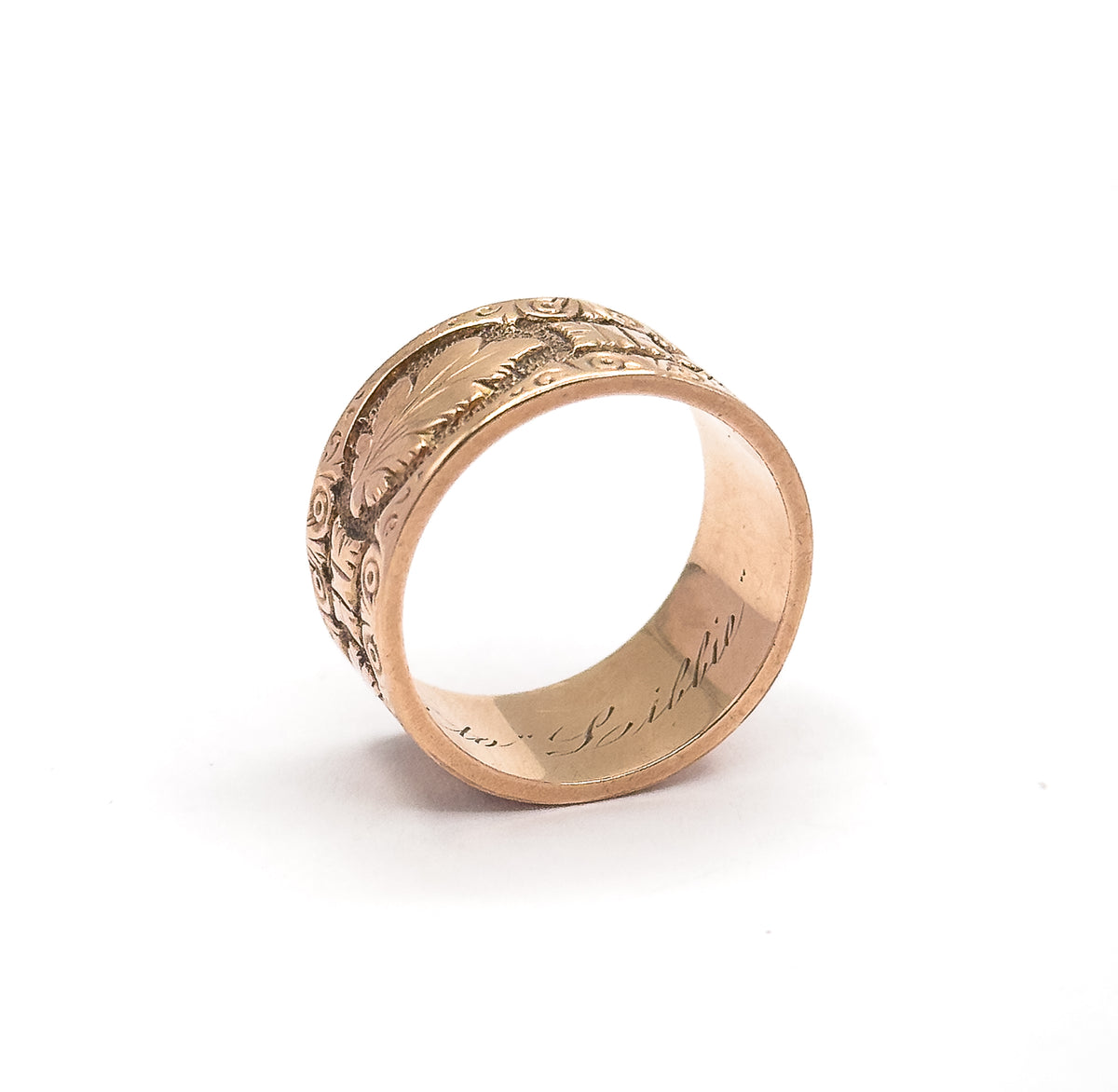 Standing upright on a plain white background, the band angled so the hand-engraved script inscription inside the shank is visible along with the leaf-covered outer surface.
