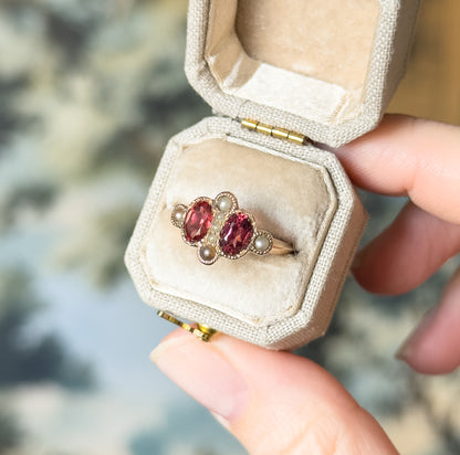 Victorian garnet and seed pearl ring in 10K rose gold, its two oval garnets ringed by seed pearls, shown nestled in a small cream ring box held between finger and thumb for scale.