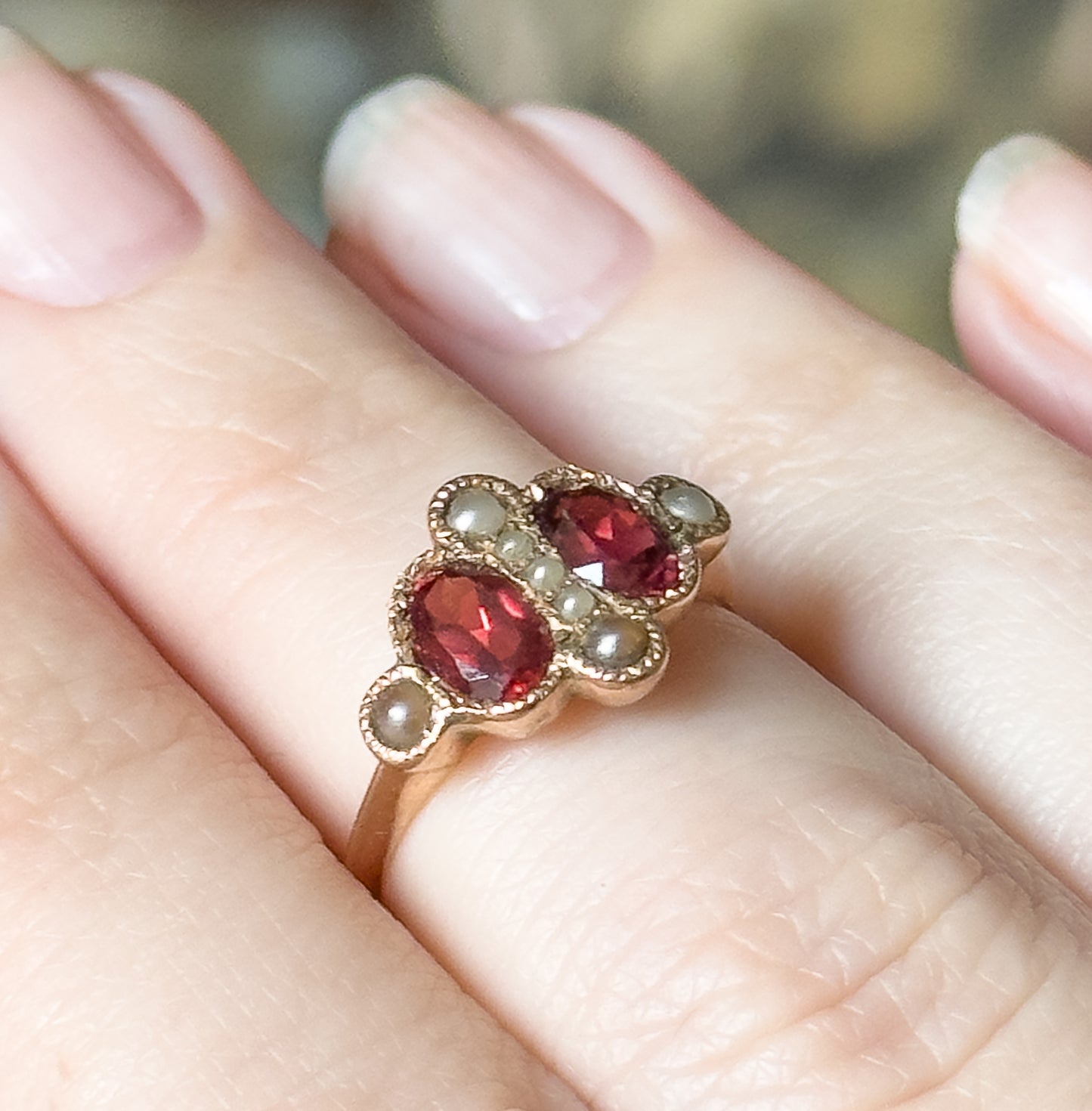 Shown on a finger in close-up, the two oval garnets sitting diagonally between seed pearls, with the rose gold face tapering low into a slender band.