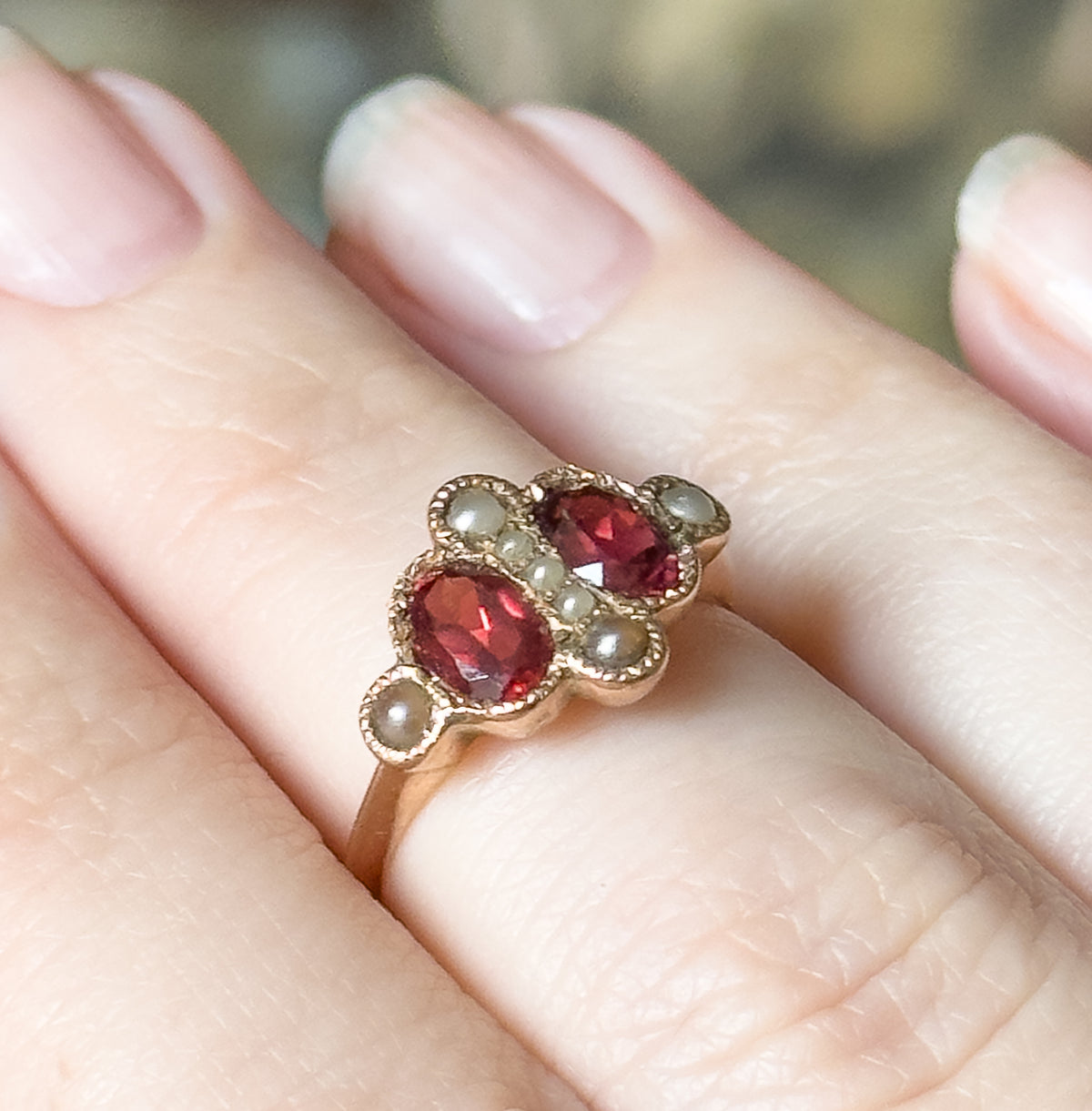 Shown on a finger in close-up, the two oval garnets sitting diagonally between seed pearls, with the rose gold face tapering low into a slender band.