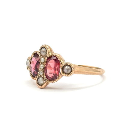 Angled three-quarter view where both oval garnets glow pink side by side, the central row of seed pearls between them and a single pearl in its milled shoulder bezel before the shank tapers away.