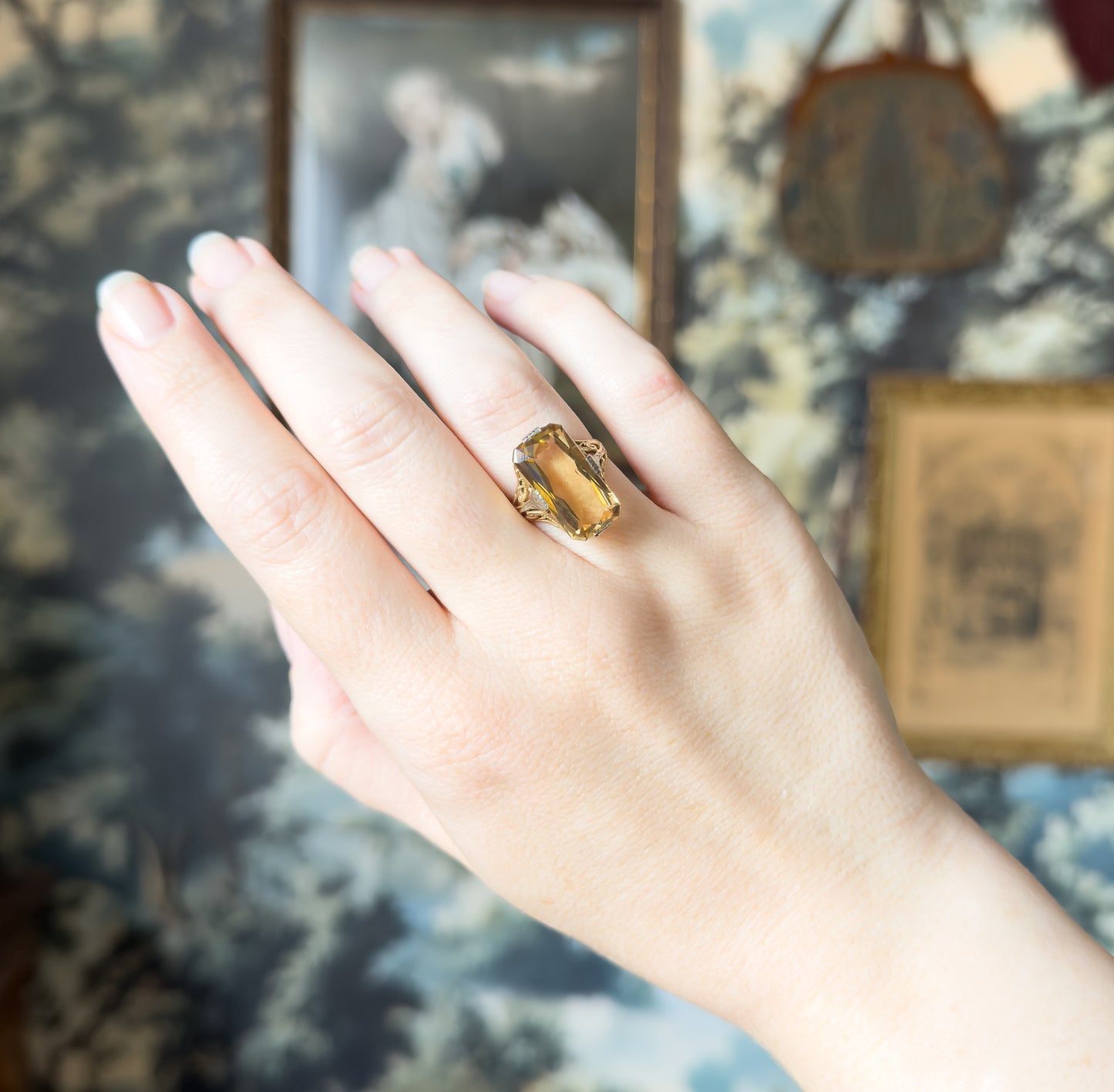 Art Deco citrine filigree ring in 14K yellow gold, shown on a finger with the elongated rectangular stone upright in its filigree setting against a soft floral backdrop.