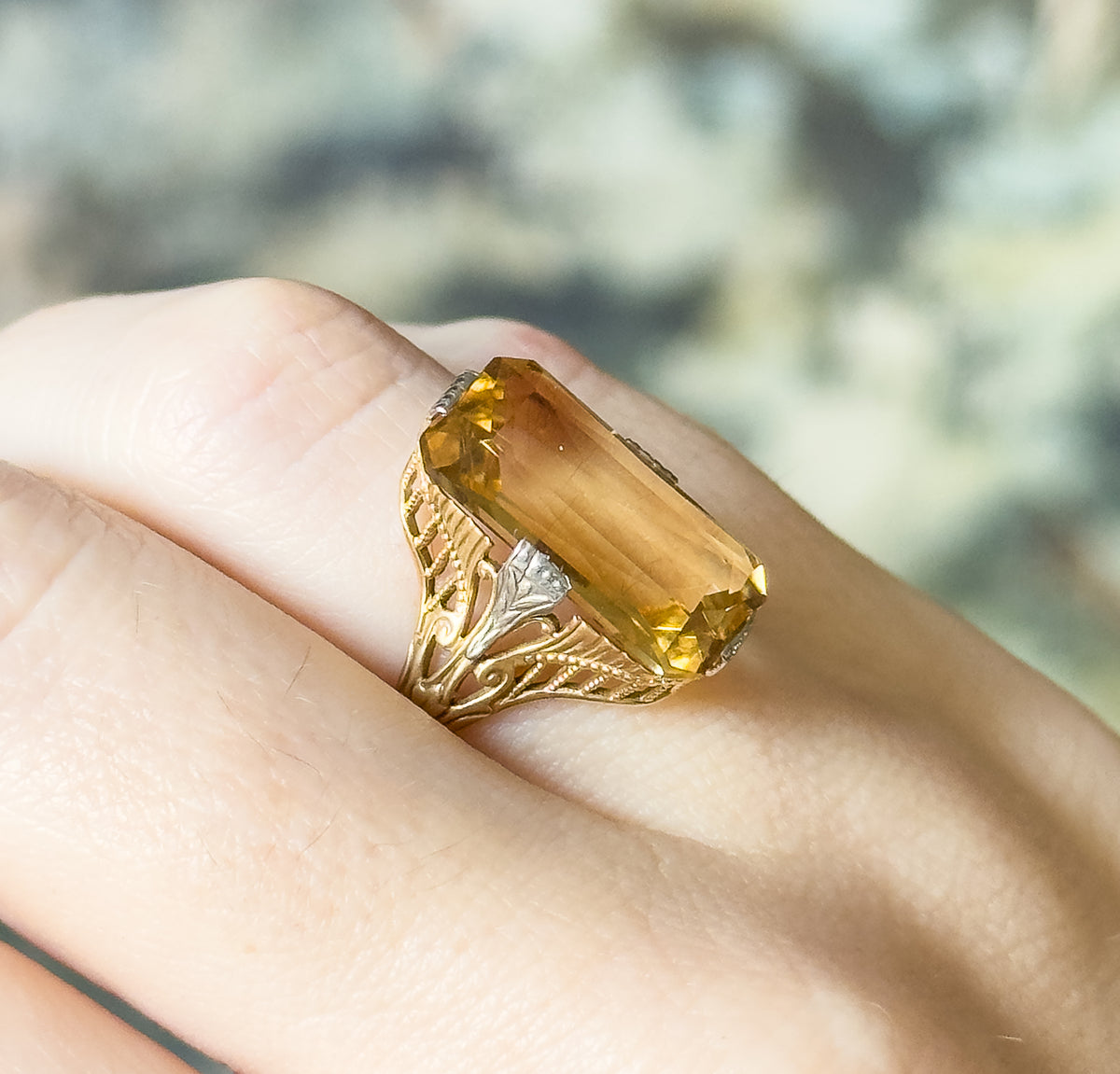 Angled top view shown on a finger, the pale orange-yellow stone lying diagonally across the knuckle so its long facets and both filigree shoulders are visible.